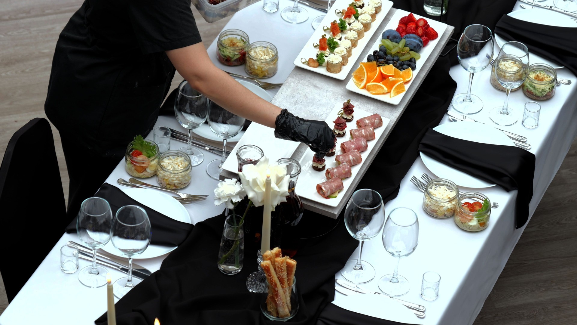 Waiter wearing black gloves arranging a variety of appetizers on an elegantly set table with a white tablecloth, black napkins, and candlelit decorations for a formal event